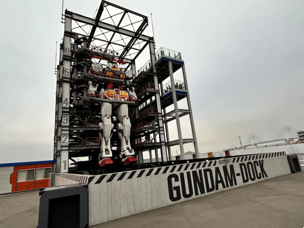 The life-sized Gundam robot at Gundam Factory Yokohama, partially enclosed in its docking station with numbered platforms and observation decks where visitors watch. The towering mecha, inspired by the iconic Mobile Suit Gundam series, stands against an overcast sky, adding to its futuristic presence at the Yokohama waterfront. © Wine Travel and Song.