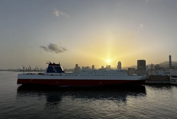A large ferry with red hull sits in Kobe port as the sun sets 
