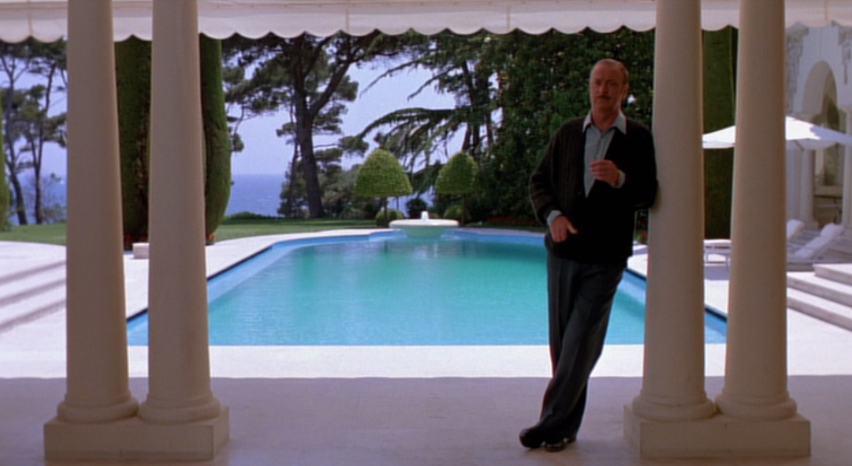 Michael Caine relaxes on a pillar in front of a pool with the Cote D'Azur coastline stretching behind him