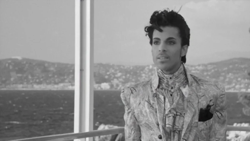 Prince, in Black and White stands on a balcony with the Baie des Anges behind him