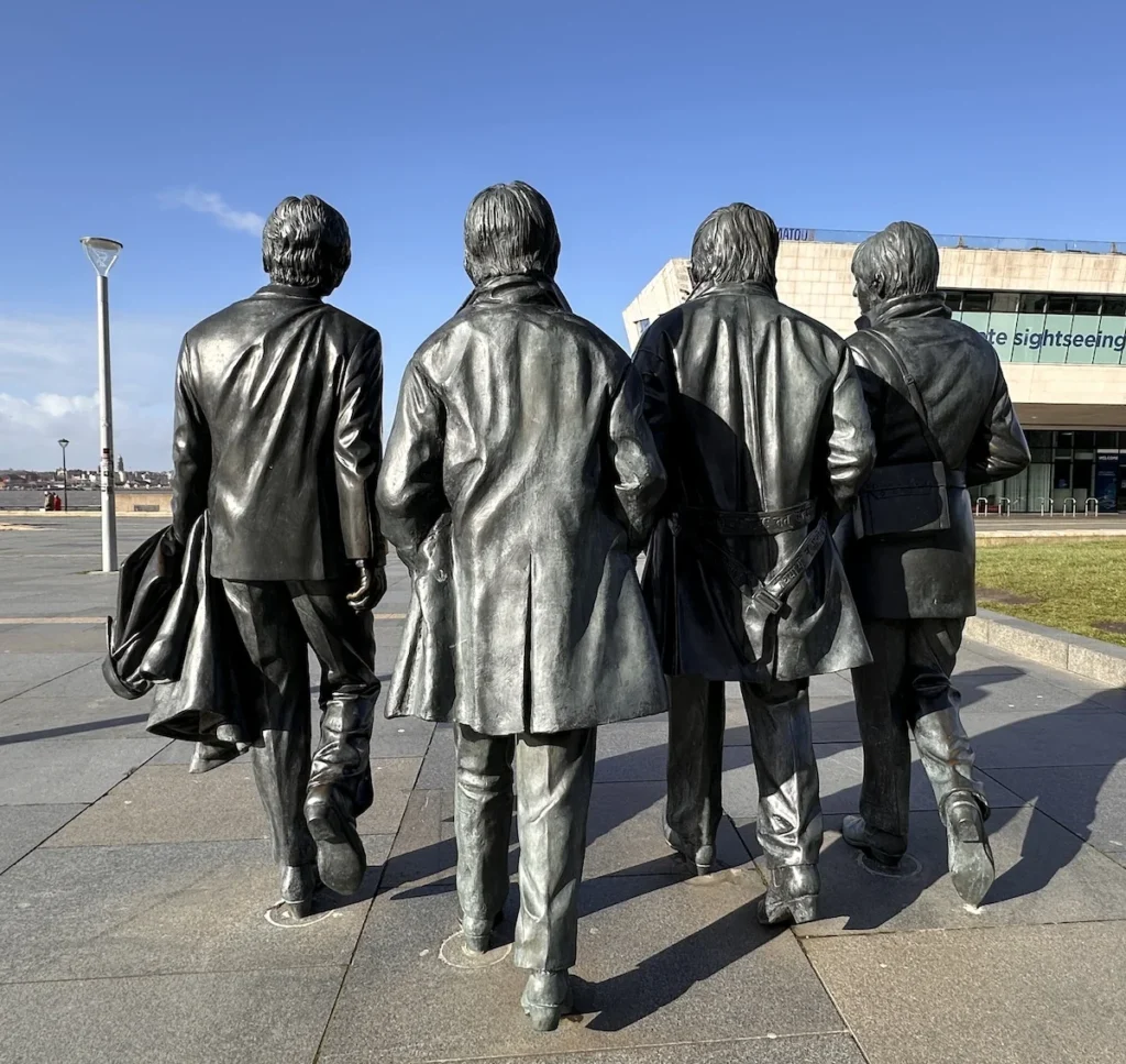 A picture of the Beatles Statue from behind looking towards Blue skies with the edge of the Mersey Ferries Building on the right hand side