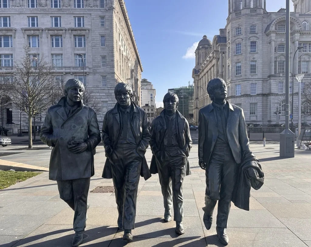 The Beatles Statue, taken from the front as if the band was walking towards you. Blue Skies frame the corner of the Liver Building behind.