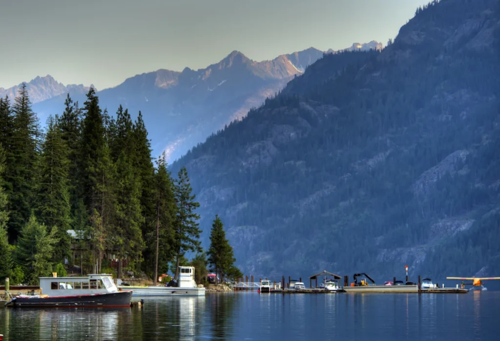 A calm lake Chelan with small boats in front of pine trees with vast mountains stretching out in the background