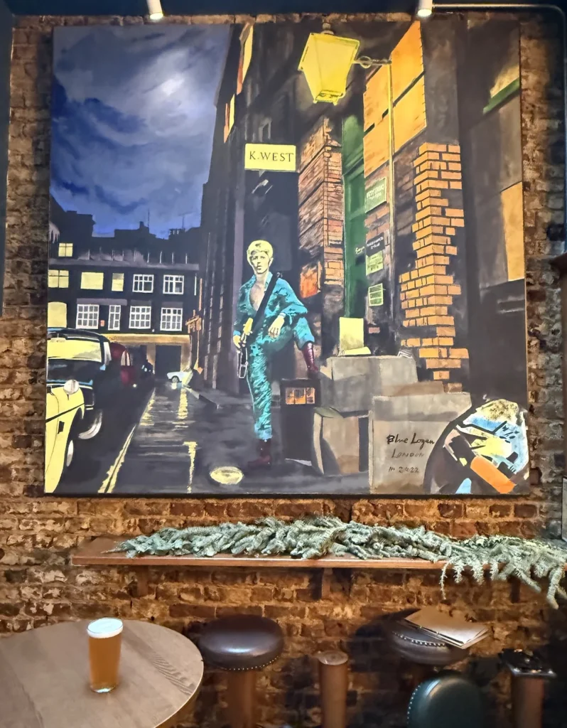 A large painting of the famous David Bowie cover hangs on a bare brick wall at the Starman pub. A pint of non alcoholic beer stands on a table in front of the artwork