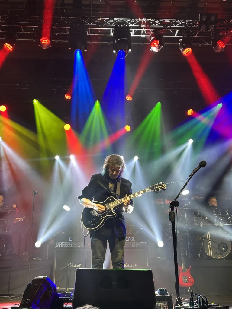 Steve Hackett wearing a black jacket playing a black guitar surrounded by lights