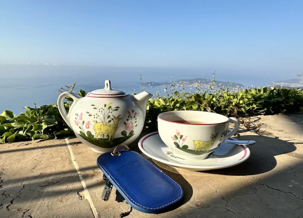 A delicate cup and Saucer with Golden Goat (Chèvre D'Or) motifs sits behind a dark blue key fob with the sea in the distance