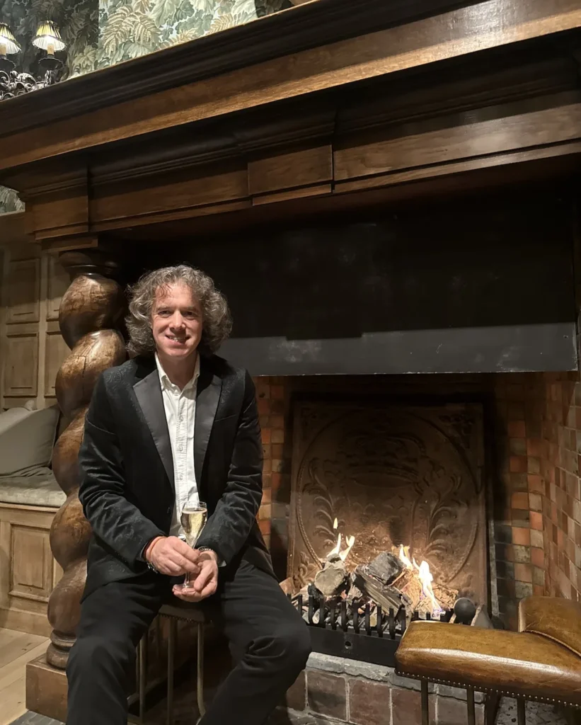 The author sits in a black silk lapel jacket and white shirt holding a glass of champagne sitting on a fender next to a roaring log fire