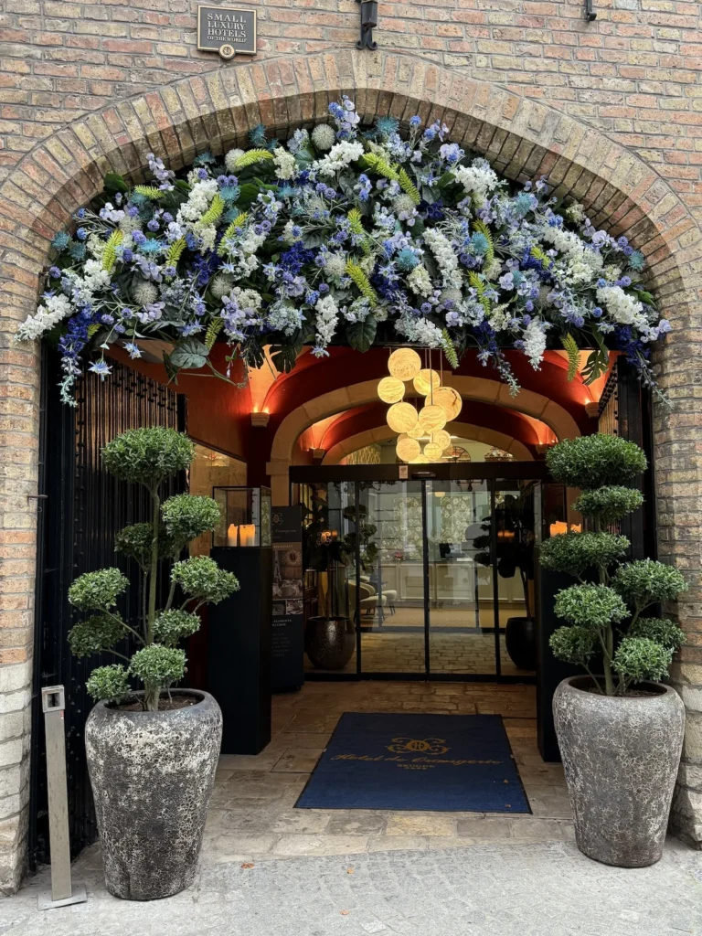 The glass entrance to the luxury hotel Orangerie is framed by two green trees and a garland of white, purple and green flowers above the door 