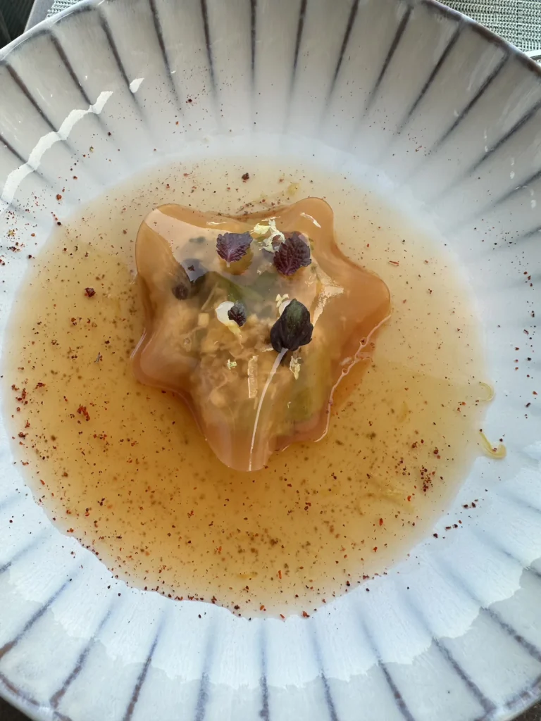 A translucent ravioli sits like a star in orange coloured juice on a white scalloped plate