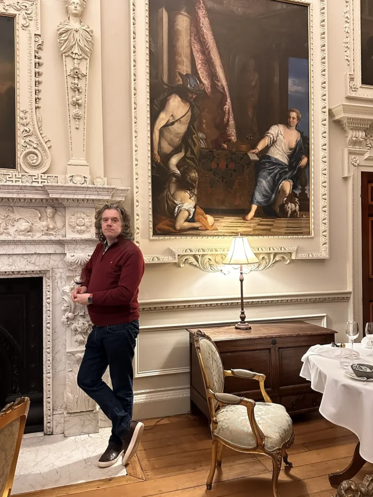 The author stands with a glass of red wine next to a large fireplace in the dining room of the luxurious Hawkstone Hall Hotel 