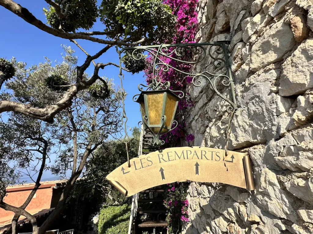 A gold sign with arrows to 'Les Remparts' Restaurant stands below a golfern lantern and pink flowers against as bright blue sky 