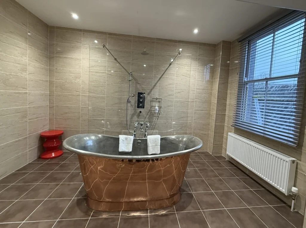 A copper freestanding bath tub stands in the middle of a large bathroom with a modern red still in the back left corner 