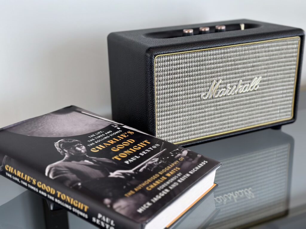 A Marshall Bluetooth Speaker sits reflected on a glass table. In the foreground is a hard copy of the Charlie Watts biography, 'Charlie's Good Tonight.'