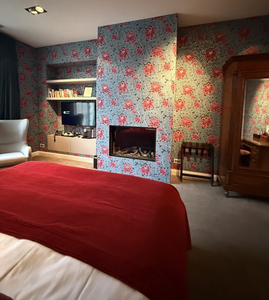 A bright red bed cover stands in the foreground against a pretty blue wallpaper with pink flowers surrounding a glass fronted gas fire in this hotel bedroom