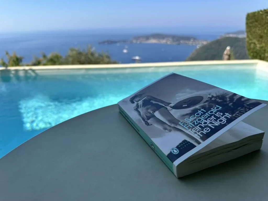 A paperback copy of 'Tender is the Night' sits on a white table in shade against the turquoise blue swimming pool and deeper blue sea behind