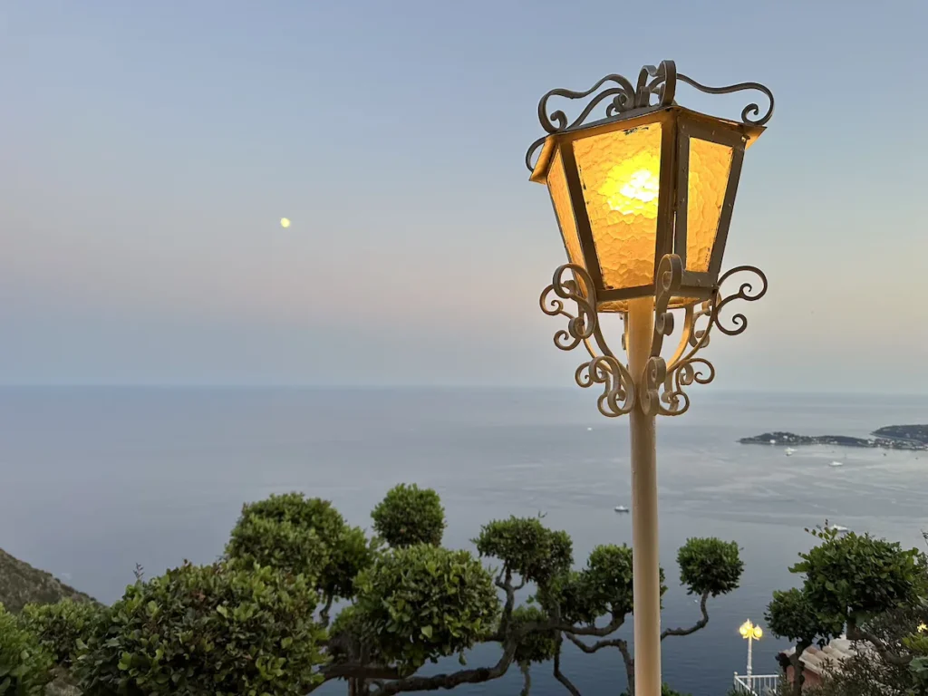 An iconic lantern offers a warm yellow light in the foreground whilst green trees frame the dark blue sea when looking out from Les Remparts restaurant in Eze