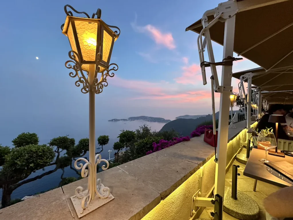 A white lamp with yellow light in the foregeround sits in front of a pastel blue sky with pink whispy clouds and the luxury Chèvre D'Or hotel.