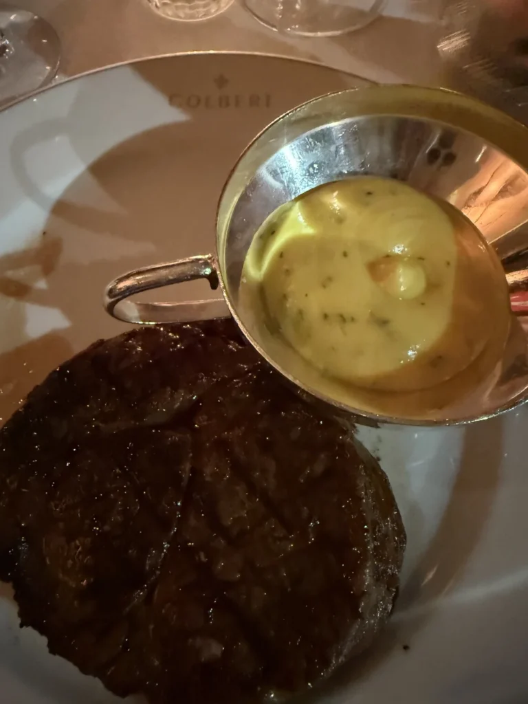 A thick steak sits upon a white Colbert crested plate with a silver dish of yellow Bernaise Sauce next to it 