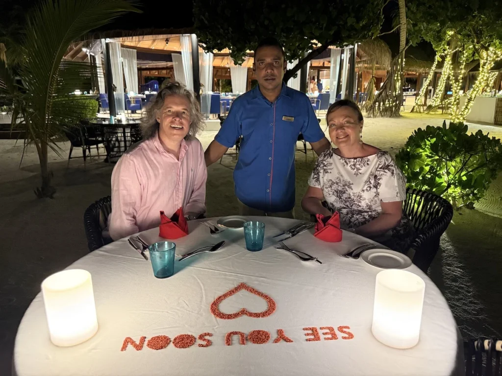 A picture of the author and his beautiful wife with Gasim our waiter standing between us. On the table in front is the phrase see you soon soon.