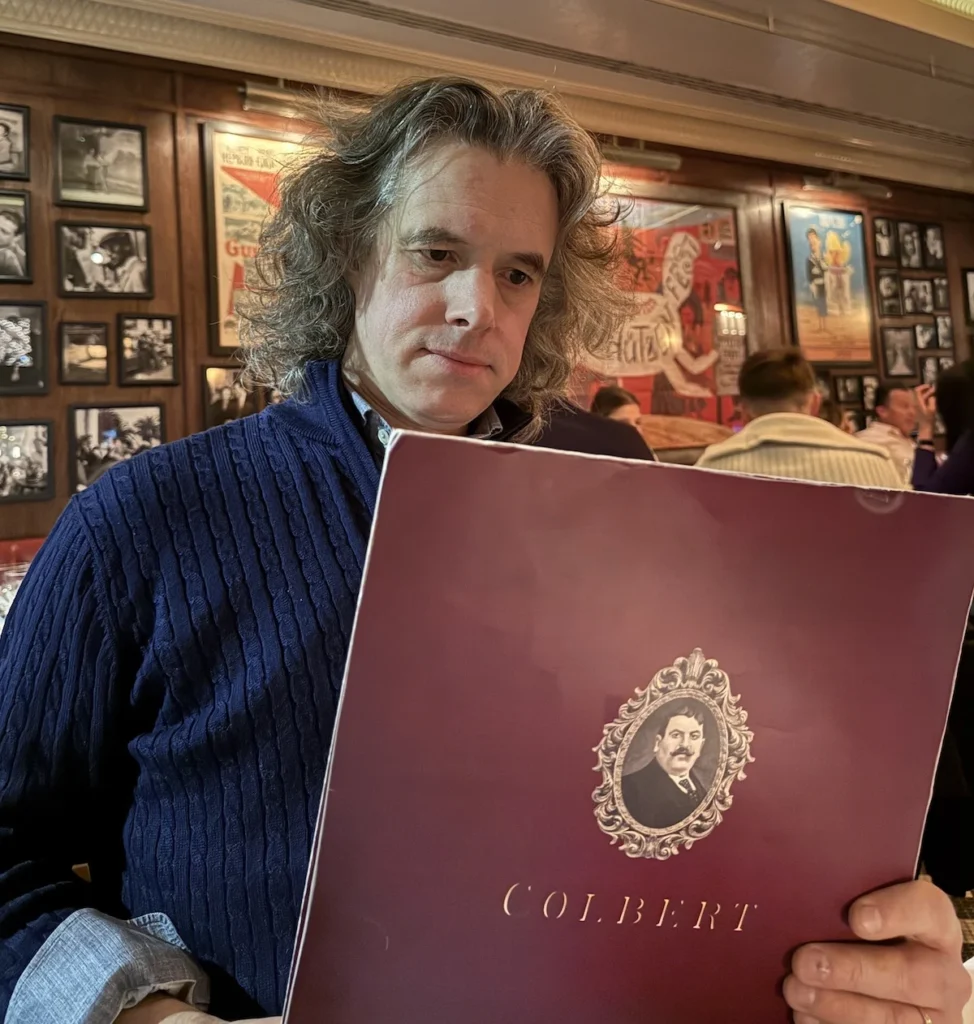 The author, Jay, wears a blue jumper with distinctive long hair, looking sat the dark red Colbert Menu