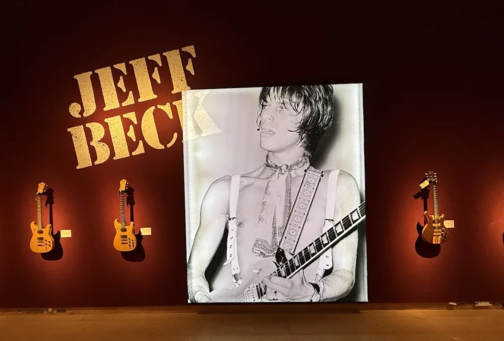 On a red wall hangs four guitars two either side of a large black and white photograph and the words JEFF BECK at Christies Auction House