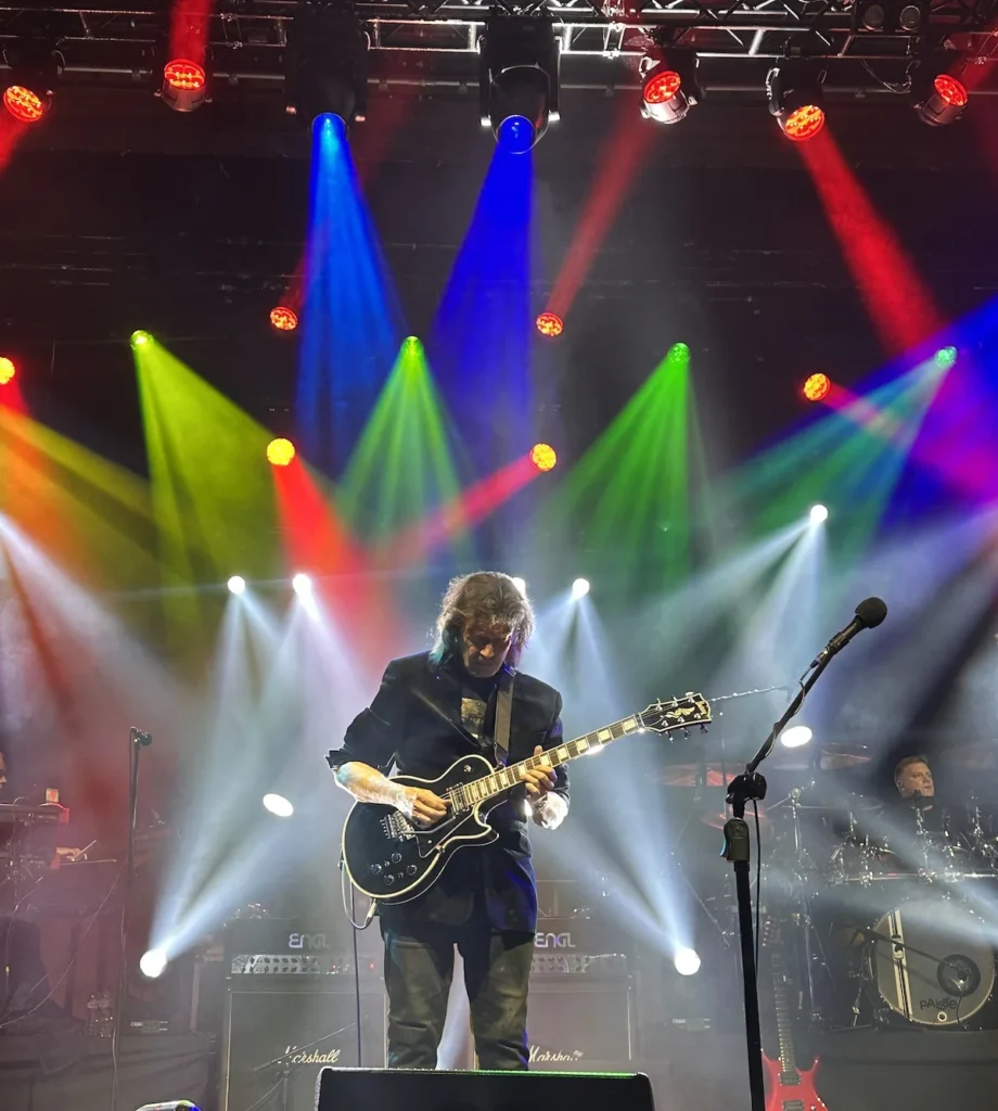 Guitarist Steve Hackett performs solo beneath red, green, white and blue spotlights, on stage in Cambridge UK. 2024