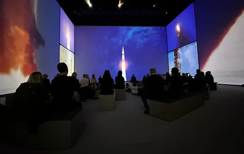 An audience, photographed from behind are dwarfed by the size of the screen surrounded them as an Apollo Mission rocket lifts off into space