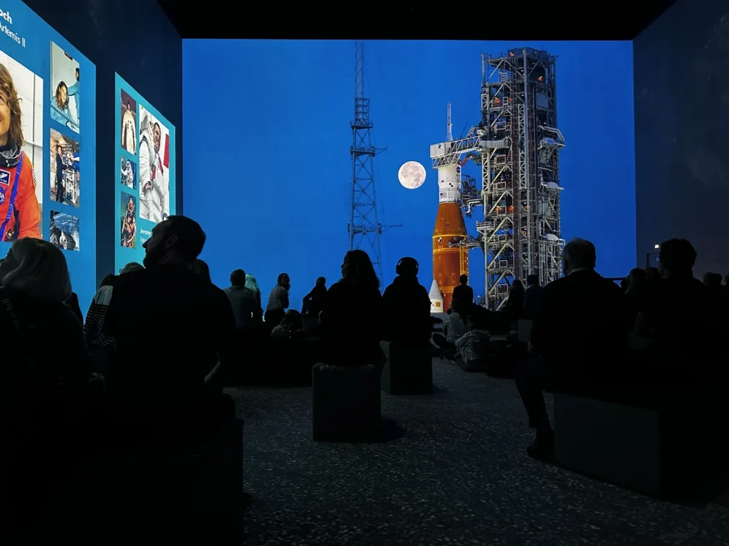 The red tip of Artemis 1 rocket stands against a darkening blue sky as the Lightoom audience in the foreground look on