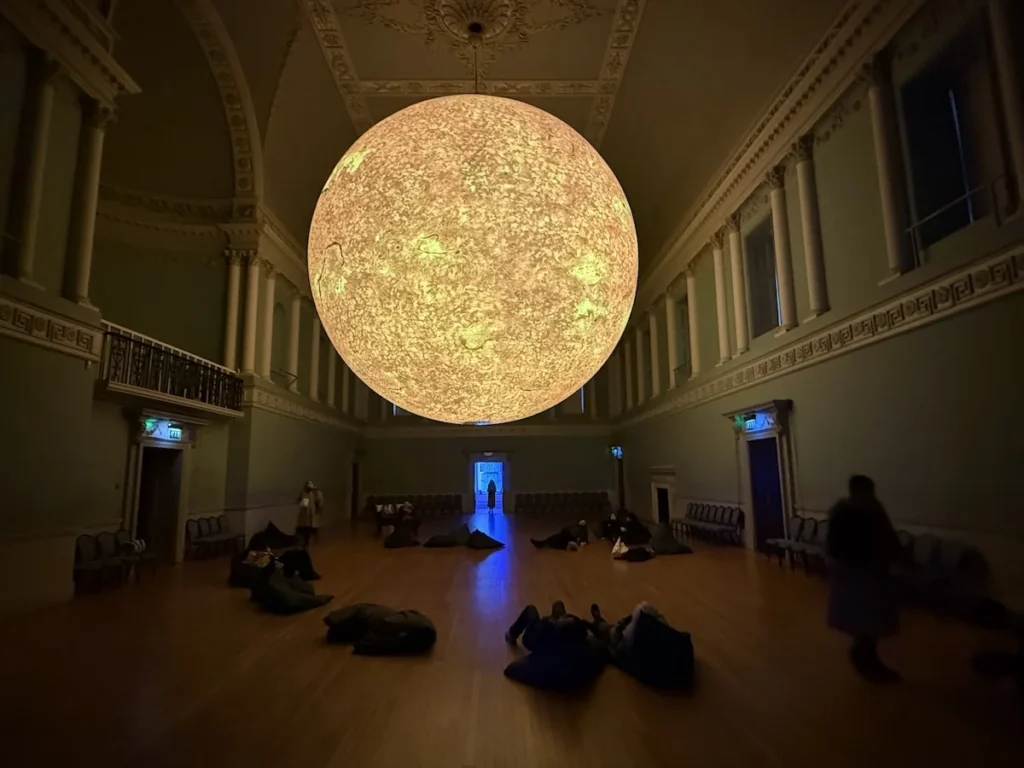 A large sun hangs in the Bath Assembly Rooms with people lying silhouetted on the floor. The yellow sun contrasts with a slip of blue light at the far end of the room