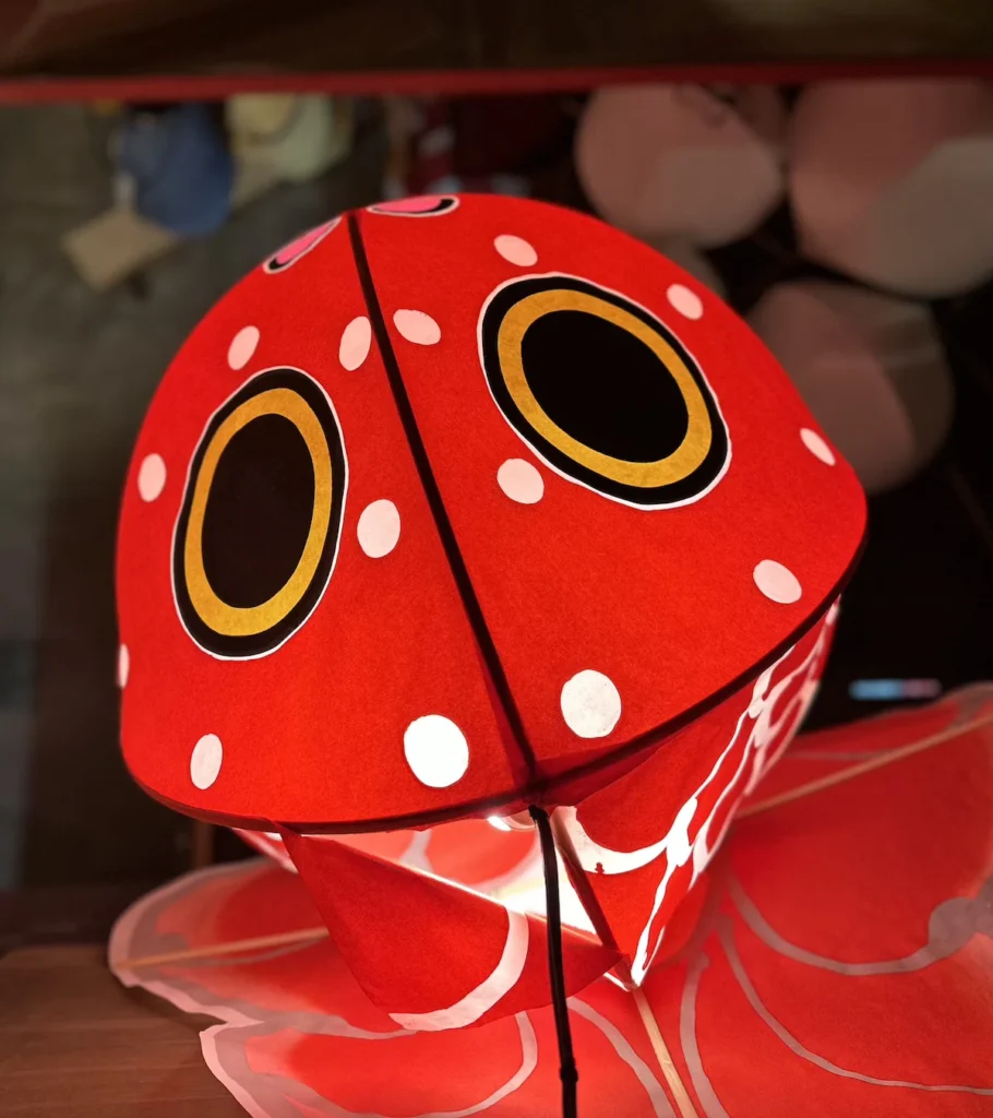 A bright red prayer lantern in Hirosaki, Japan, shaped like a traditional festival decoration with large, circular eyes and white polka dots. The glowing interior casts a warm light, highlighting its intricate hand-painted details. © Wine Travel and Song.
