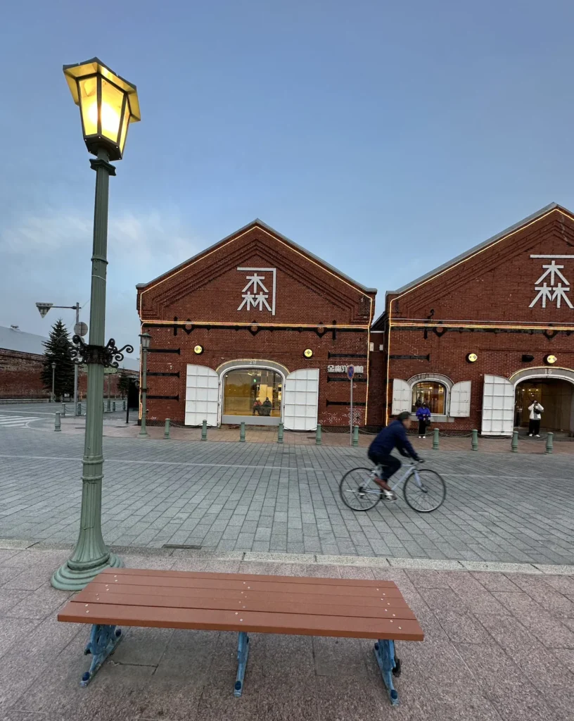 A cyclist rides past the historic red brick warehouse in Hakodate, Japan, with a bench and street lamp in the foreground.