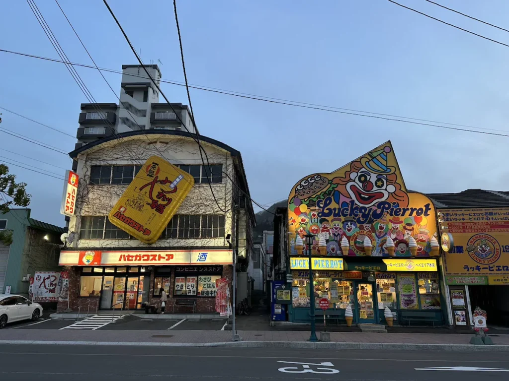 A lively street scene in Hakodate, Japan, featuring a mix of traditional wooden storefronts and modern shops under an overcast sky.