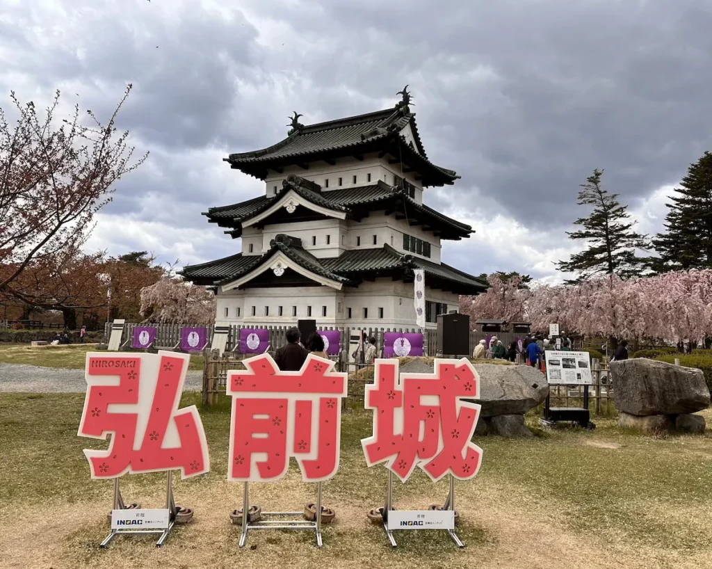 Hirosaki Castle in Aomori, Japan, a historic three-story keep with traditional black-tiled roofs and white walls, surrounded by cherry blossoms. In the foreground, a large red and white sign with ‘Hirosaki Castle’ (弘前城) stands as a popular photo spot. © Wine Travel and Song