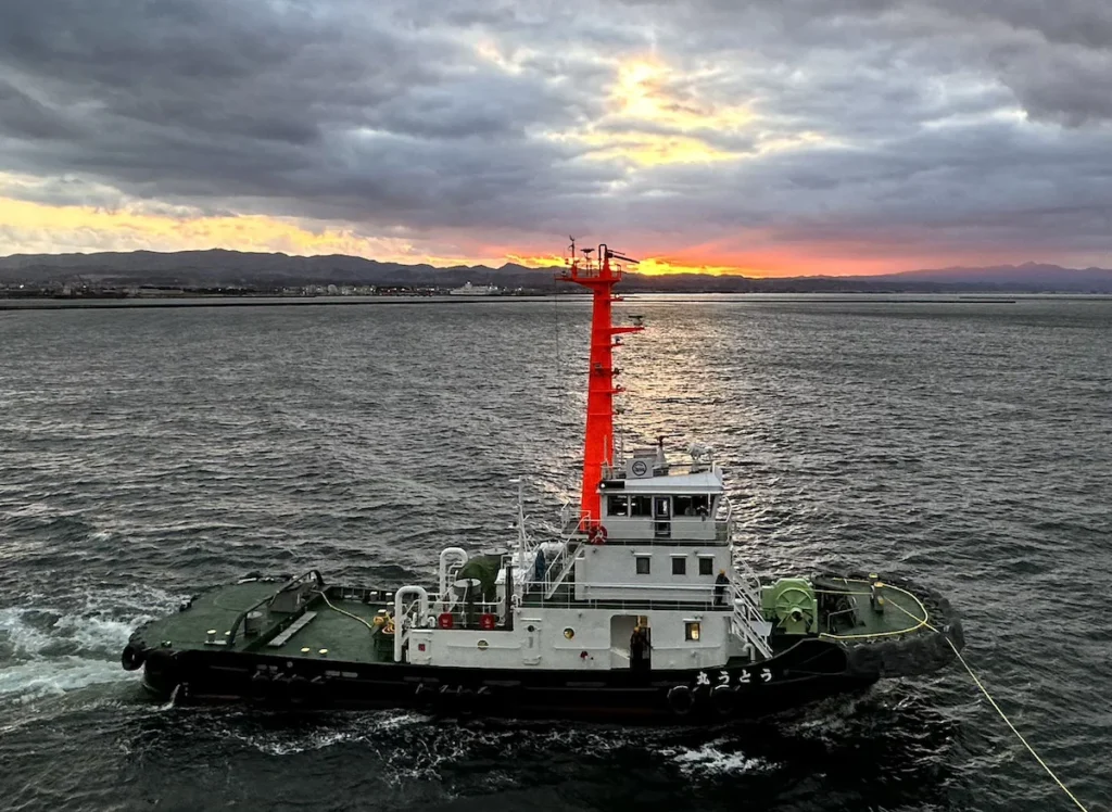 A Japanese pilot boat with a striking red mast and deep green deck sails through dark waters at sunrise. The golden-pink hues of the rising sun break through the heavy clouds, casting a dramatic glow over the horizon. A vibrant contrast of colours, reflecting the rich visual identity of Japan. © Wine Travel and Song.