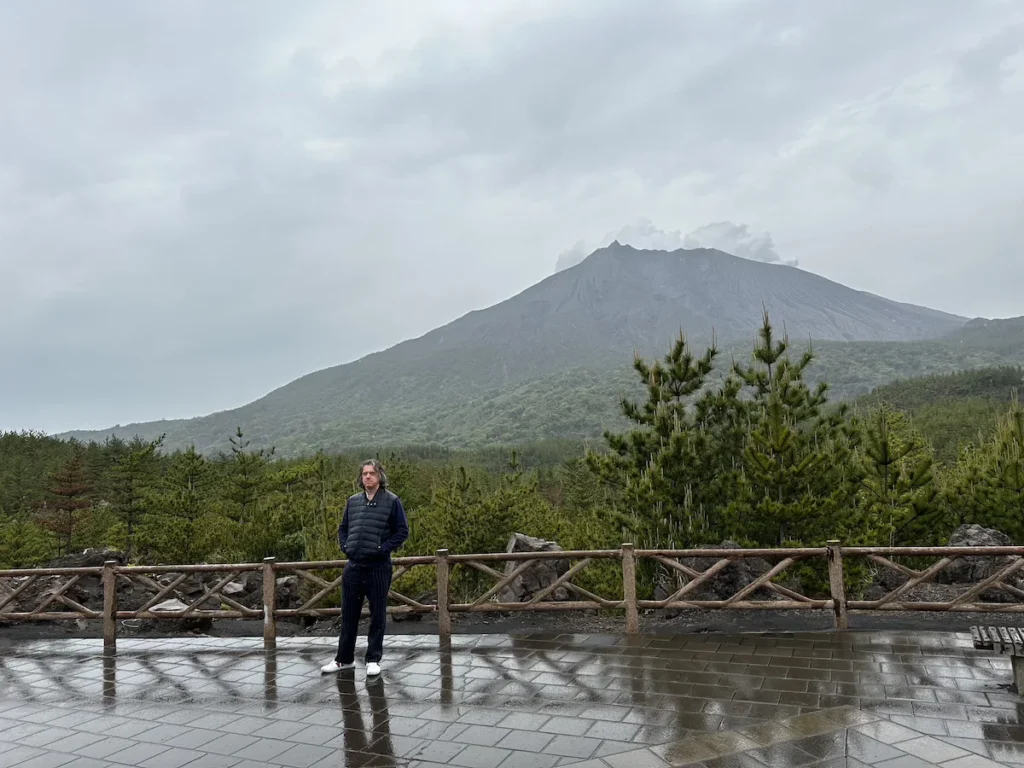 The author standing in front of Sakurajima volcano in Kagoshima, Japan, with the misty peak and rising smoke visible in the background on an overcast day."

