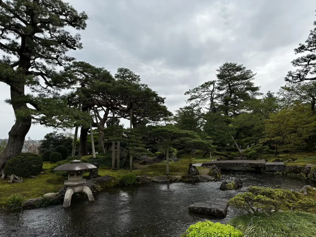 A tranquil Japanese garden in Kanazawa with sculpted pine trees, a stone lantern, and a reflective pond beneath an overcast sky