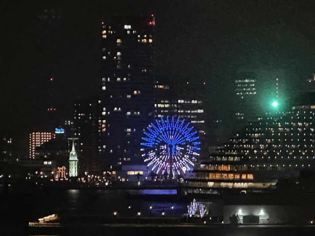 The illuminated Ferris wheel at Kobe Harborland stands against the city skyline at night, reflecting on the calm waters of Kobe Port. The vibrant lights add a dynamic contrast to the modern waterfront, a popular destination for visitors. © Wine Travel and Song.
