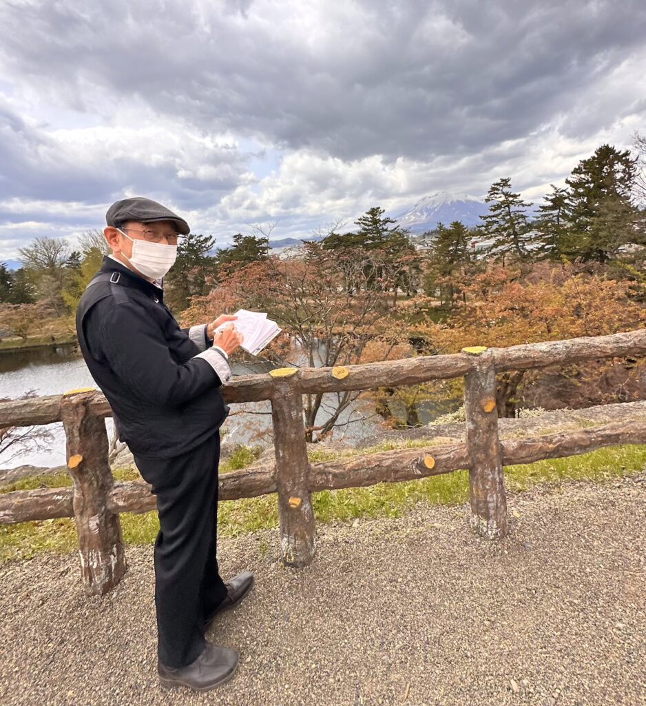 A local artist wearing a flat cap and face mask sketches the distant snow-capped Mount Iwaki from a scenic viewpoint at Hirosaki Castle, Japan. The rustic wooden fence frames the autumnal landscape, with a tranquil pond and colourful foliage in the foreground. © Wine Travel and Song.