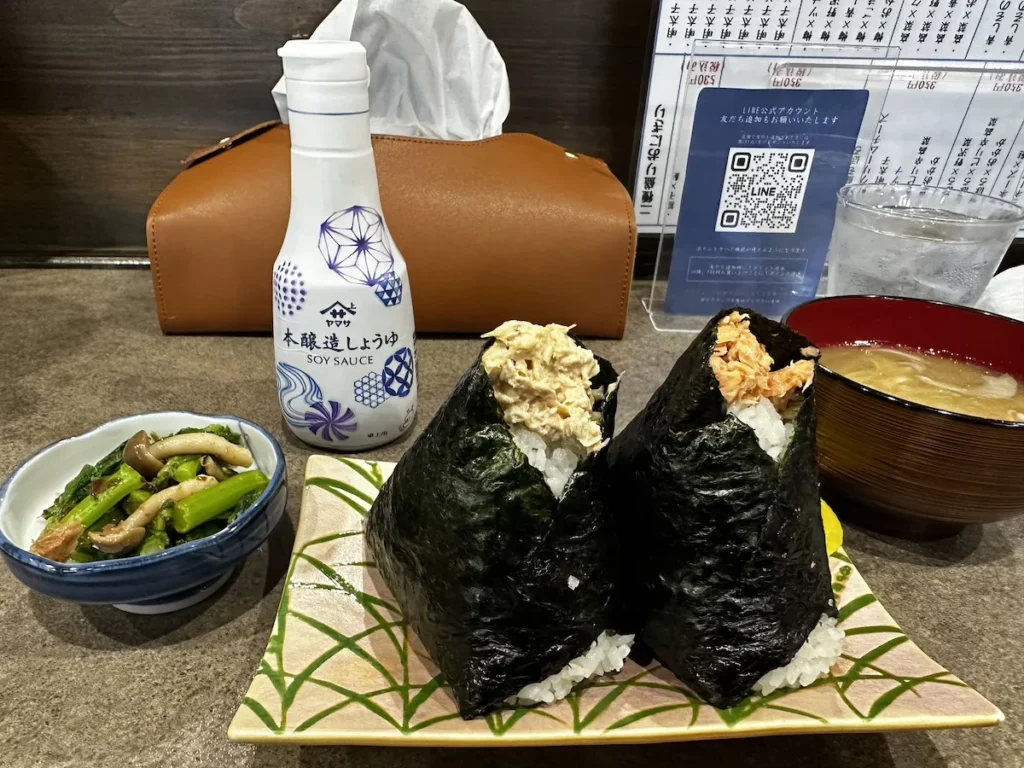 A traditional Japanese meal featuring large onigiri with salmon and tuna fillings, a bowl of miso soup, and a side of pickled greens, enjoyed at a local restaurant in Niigata