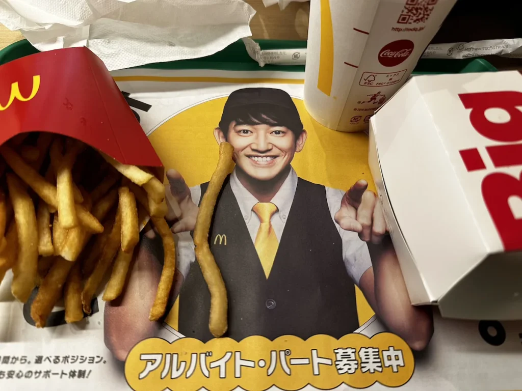 McDonald’s tray in Kanazawa, Japan, featuring a recruitment advertisement with a smiling employee in a yellow tie and black cap. A single French fry humorously aligns with his outstretched hand, blending into the image. The tray holds a red McDonald’s fries container, a soft drink cup, and a partially visible burger box. © Wine Travel and Song.