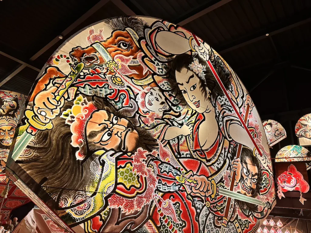 A vividly painted Nebuta-style drum in Hirosaki, Japan, featuring bold red, gold, and black designs of fierce warriors and mythical figures. The intricate brushwork and dynamic expressions capture the energy of traditional Japanese festival art. © Wine Travel and Song.