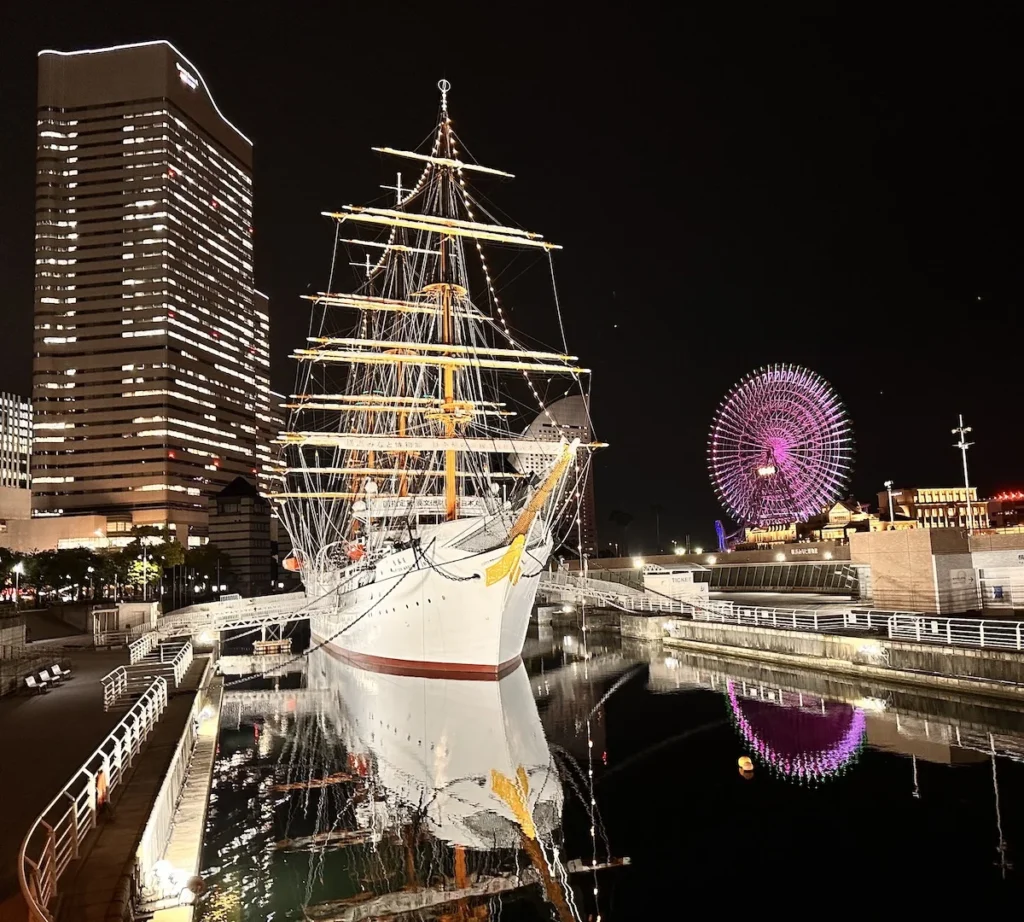 The Nippon Maru, a historic white-hulled four-masted sailing ship, illuminated at night in Yokohama’s Minato Mirai 21 district. In the background, the brightly lit Cosmo Clock 21 Ferris wheel at Yokohama Cosmo World amusement park adds to the vibrant cityscape, highlighting popular attractions for Japan cruise visitors.