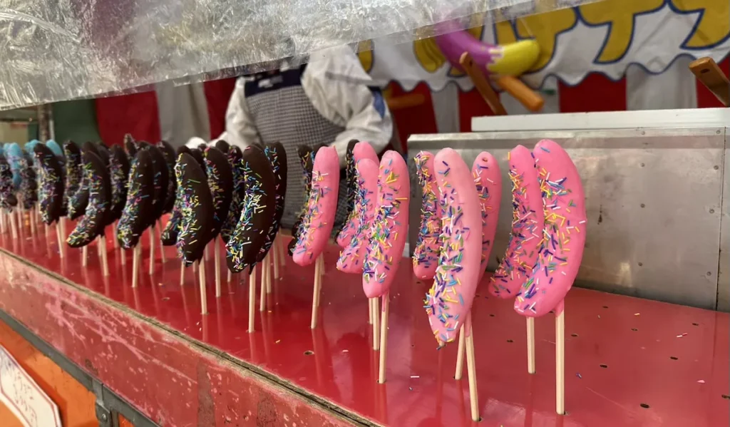 Chocolate and pink candy-coated bananas on sticks, covered in colourful sprinkles, displayed at a food truck in Aomori, Japan. A popular festival treat, these sweet snacks are a common sight at Japanese street markets and yatai stalls. © Wine Travel and Song