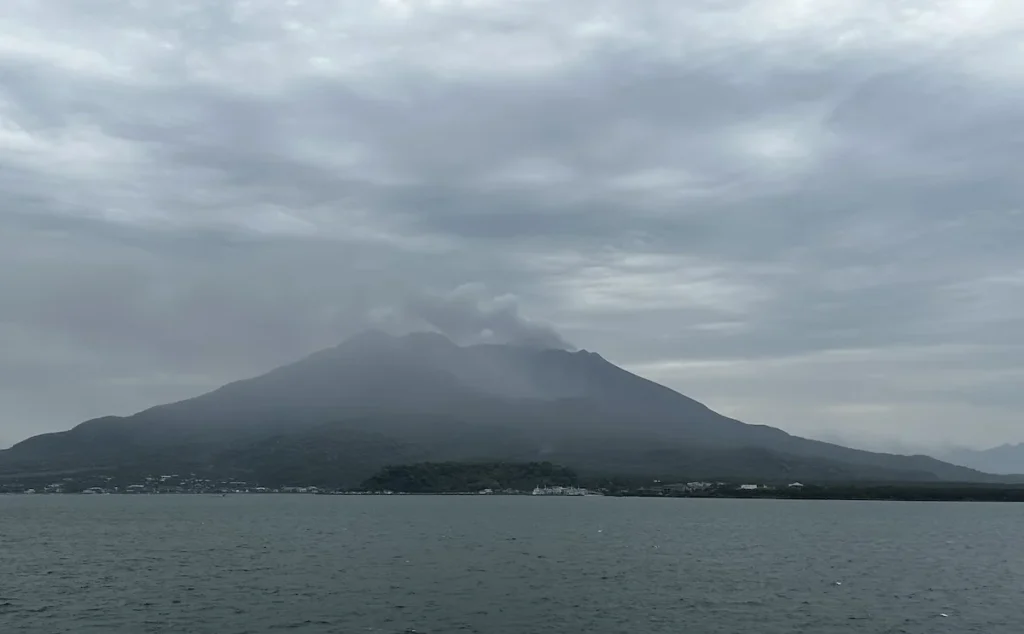 A misty view of Sakurajima volcano in Kagoshima, Japan, with smoke rising from its peak under an overcast sky, seen from across the bay.