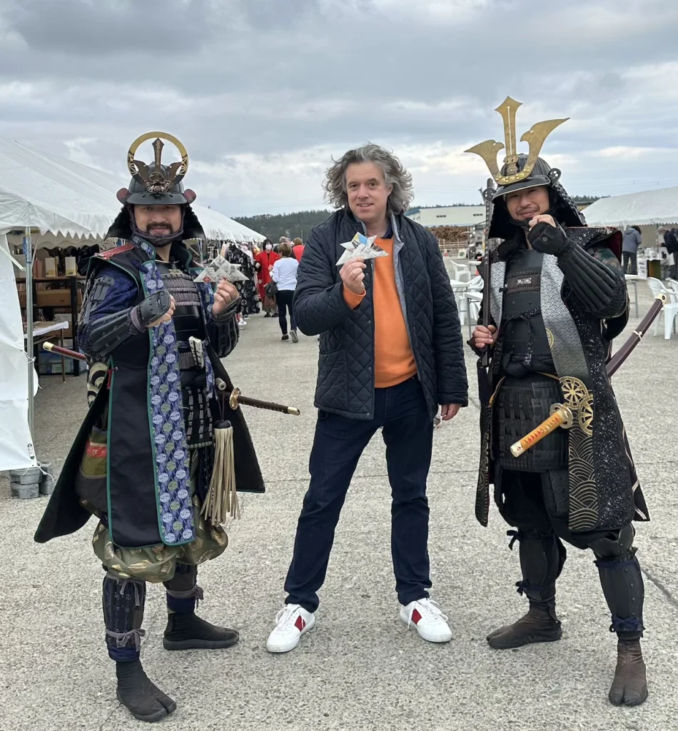 Author Jay at the Sakata greeting event in Japan, posing with two men dressed as samurai warriors in ornate traditional armour. All three hold folded paper shuriken (throwing stars), adding a playful touch to the scene. White tents and festival stalls line the background, capturing the lively atmosphere of the gathering. © Wine Travel and Song.