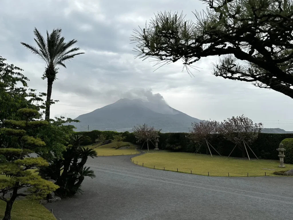 A serene Japanese garden at Sengan-en, framed by lush greenery and traditional landscaping, with the misty Sakurajima volcano looming in the background.