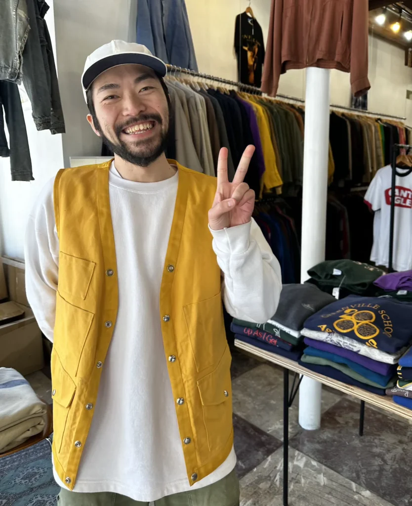 A friendly shop owner at THIS MAN vintage clothing store in Niigata, Japan, wearing a mustard yellow vest and white cap, flashing a peace sign with a warm smile. Behind him, racks of vintage t-shirts and jackets fill the stylish boutique, known for its curated retro fashion. Location: THIS MAN, 4 Bancho-568 Furumachidori, Chuo Ward, Niigata, 951-8063, Japan. © Wine Travel and Song.
