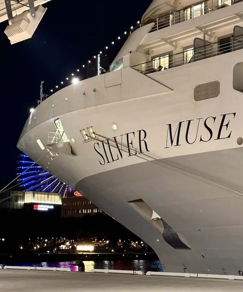 “The Silver Muse cruise ship docked at Yokohama Port at night. In the background, the brightly illuminated Ferris wheel adds a colourful glow to the skyline.