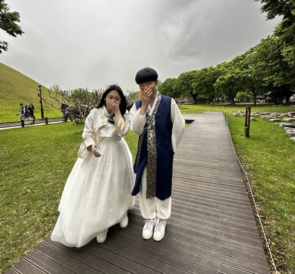 A young couple dressed in traditional hanbok share a shy smile in a park in Gyeongju