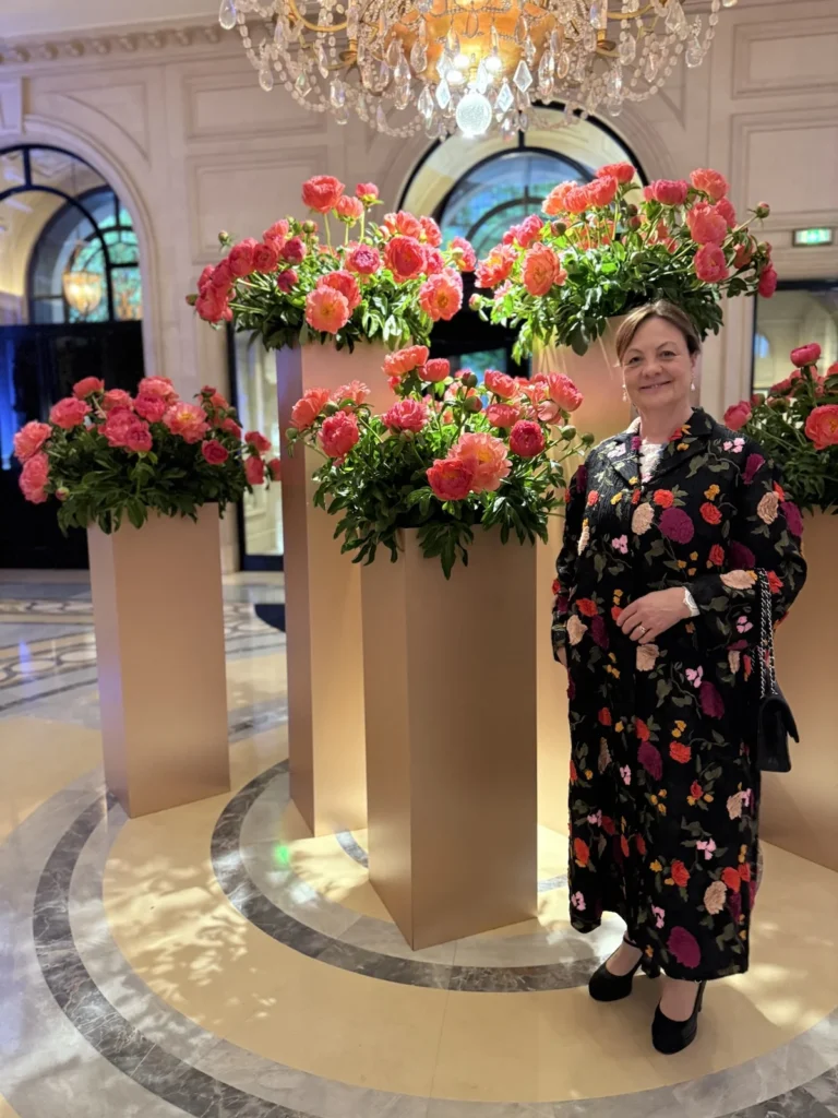 Katherine standing in the George V hotel lobby surrounded by coral and pink peonies under a chandelier.